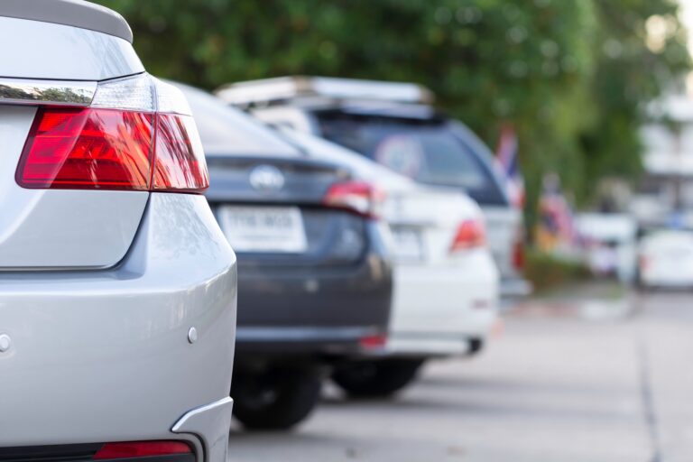 A row of modern cars parked along a street, shown in a shallow depth-of-field close-up focusing on the taillight of a silver sedan.